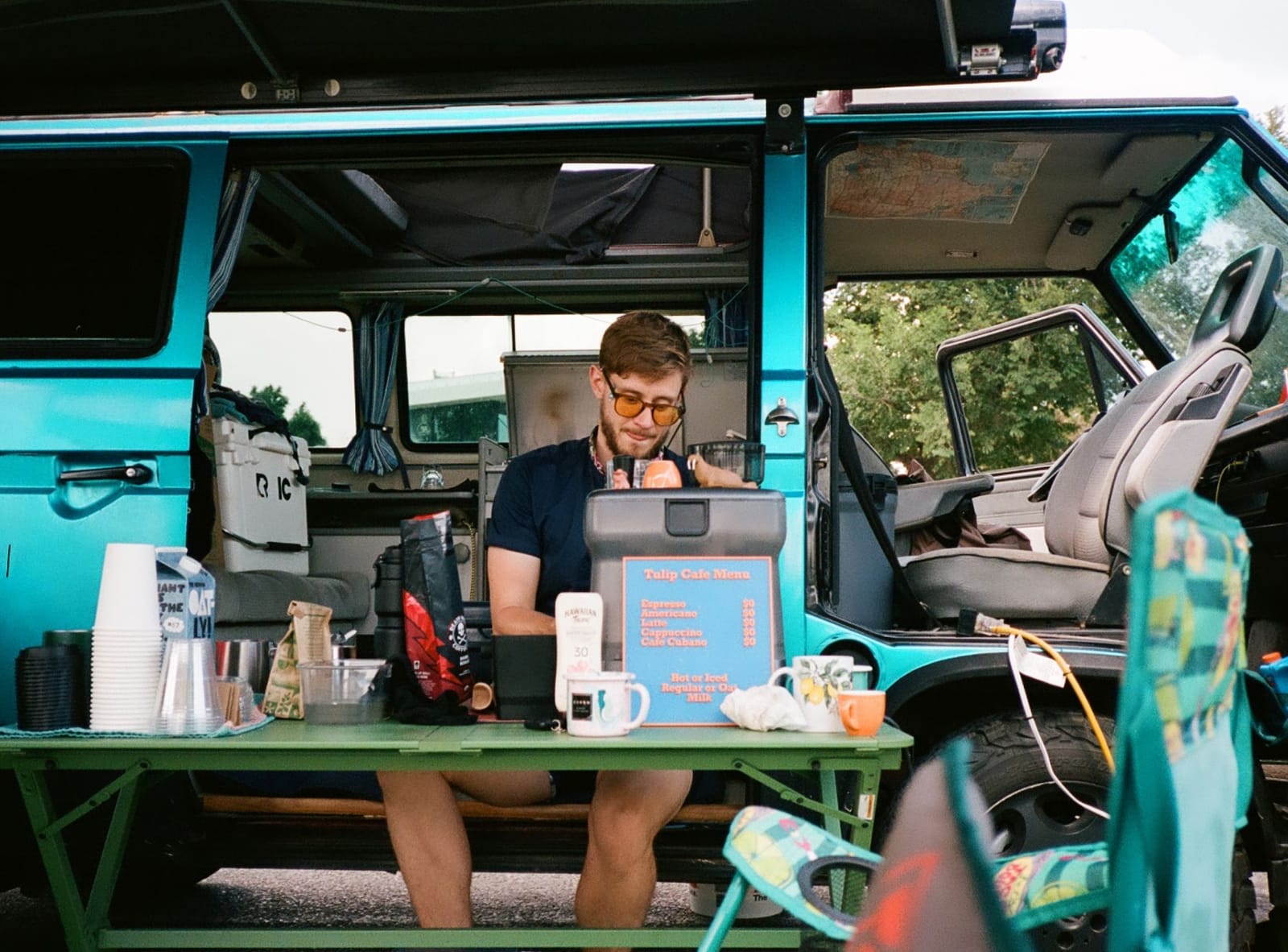 Will behind the table at the van's side door, making coffee